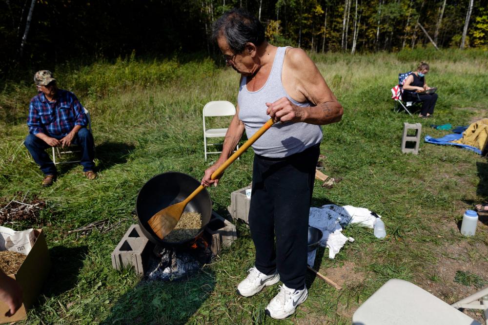 Man using long-handled instrument to stir wild rice in a large skillet with people seated nearby and forest in the background