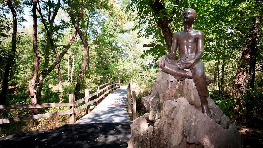 Dark-colored statue sitting atop rock to the right of a bridge cutting through stand of trees on a sunny day