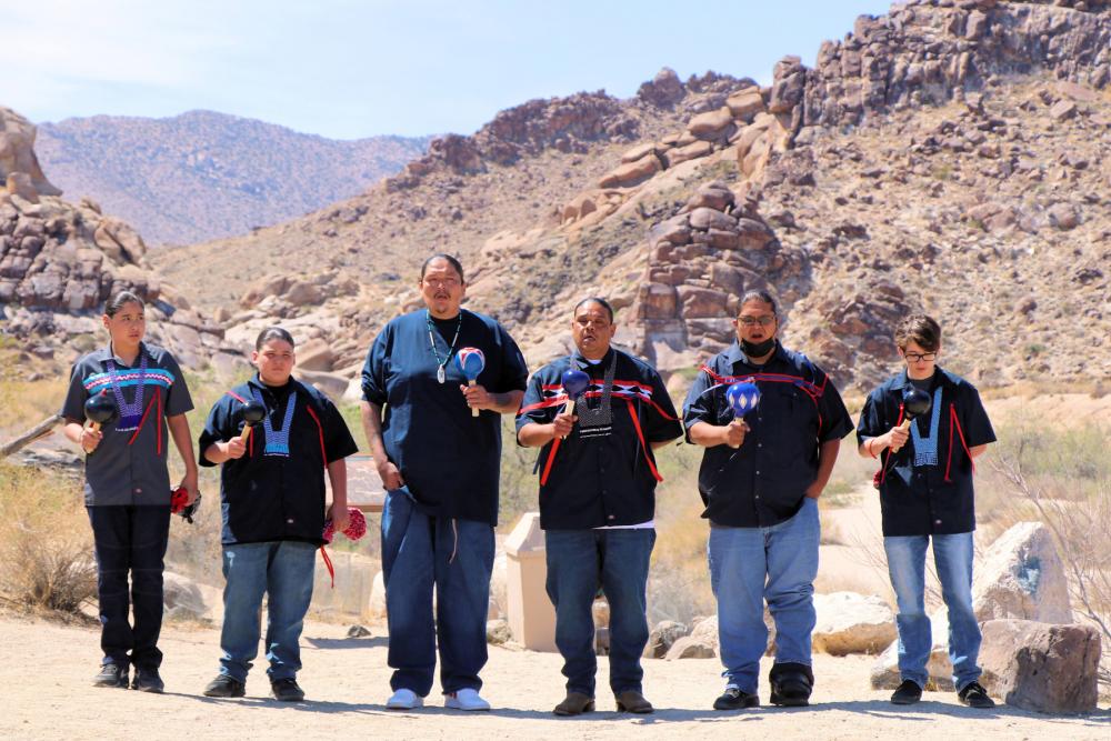 Six singers in modern Fort Mojave tribe garb playing traditional instruments in a rocky desert landscape