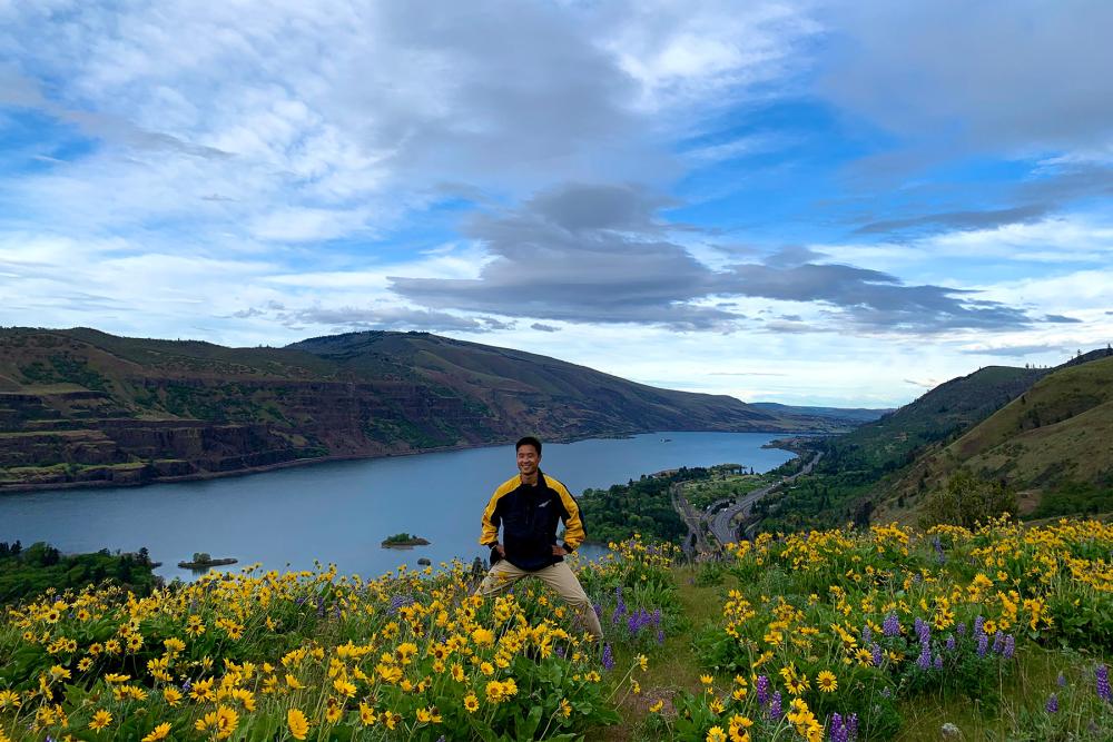 man standing in a field of flowers with a lake and mountains in the background 