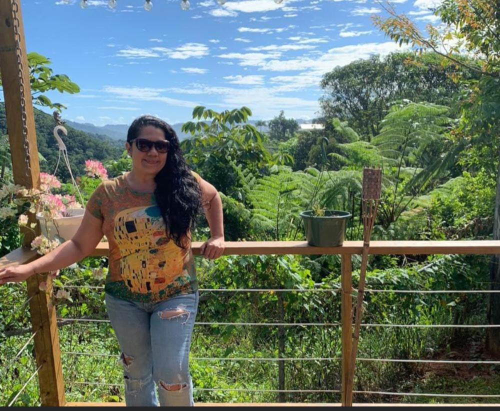 person posing on a wooden balcony with trees and tropical plants in the background