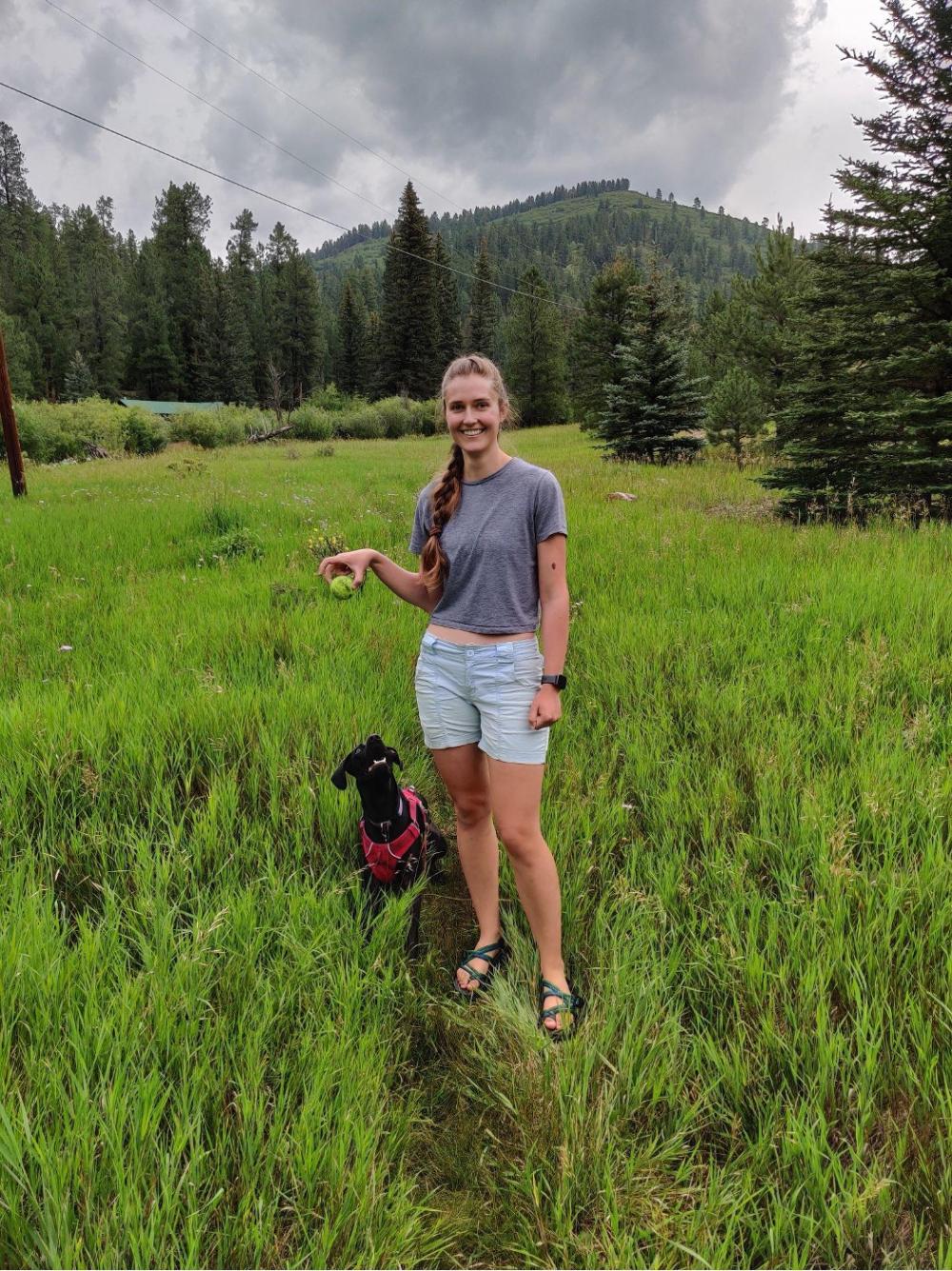 Rachel holds a tennis ball with her dog in front of a landscape.