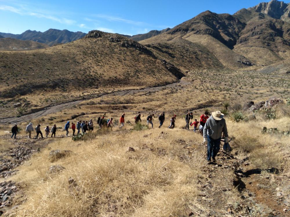 Procession of people hiking through mountainous desert landscape, with leader of the group near the foreground
