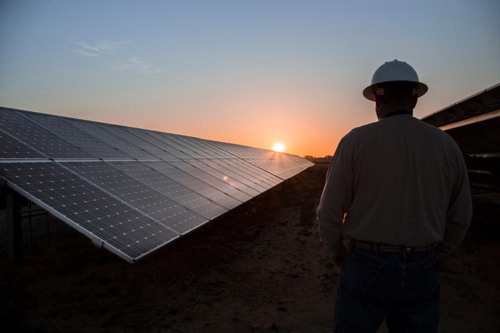 Helmet worker in the foreground looks at sunrise past long solar panel array