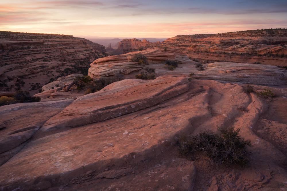 Reddish rock formations in immediate foreground and canyons and buttes in background, with sunrise behind them