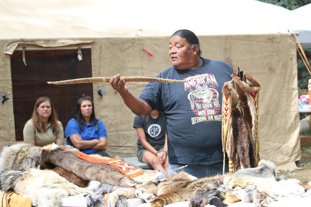 Man holds up a snake-skin covered hunting bow with three people sitting in front of a tent visible behind him