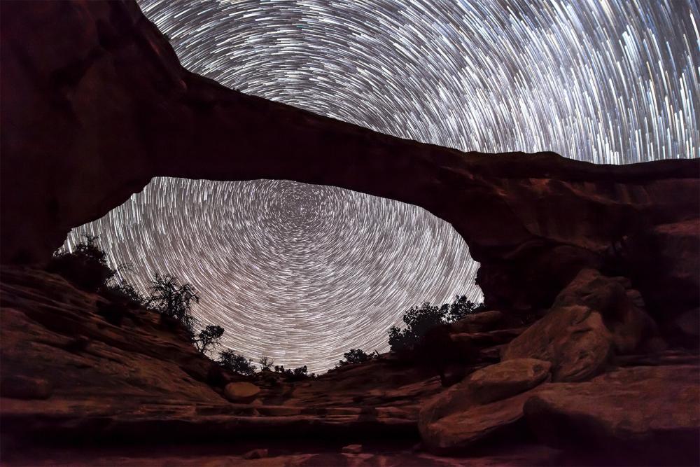 timelapse picture of stars over rock formation taht looks like a bridge