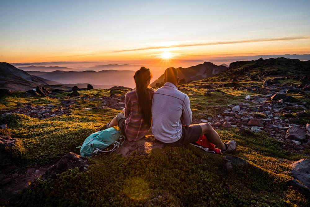 Two people sitting together on a small hill surrounded by rocky terrain, looking toward the setting sun