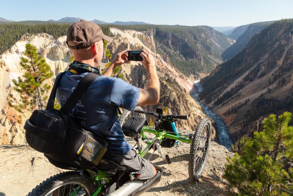 One man is on a three wheeled chair sitting at the rim of the Grand Canyon of the Yellowstone taking photos of the canyon below.