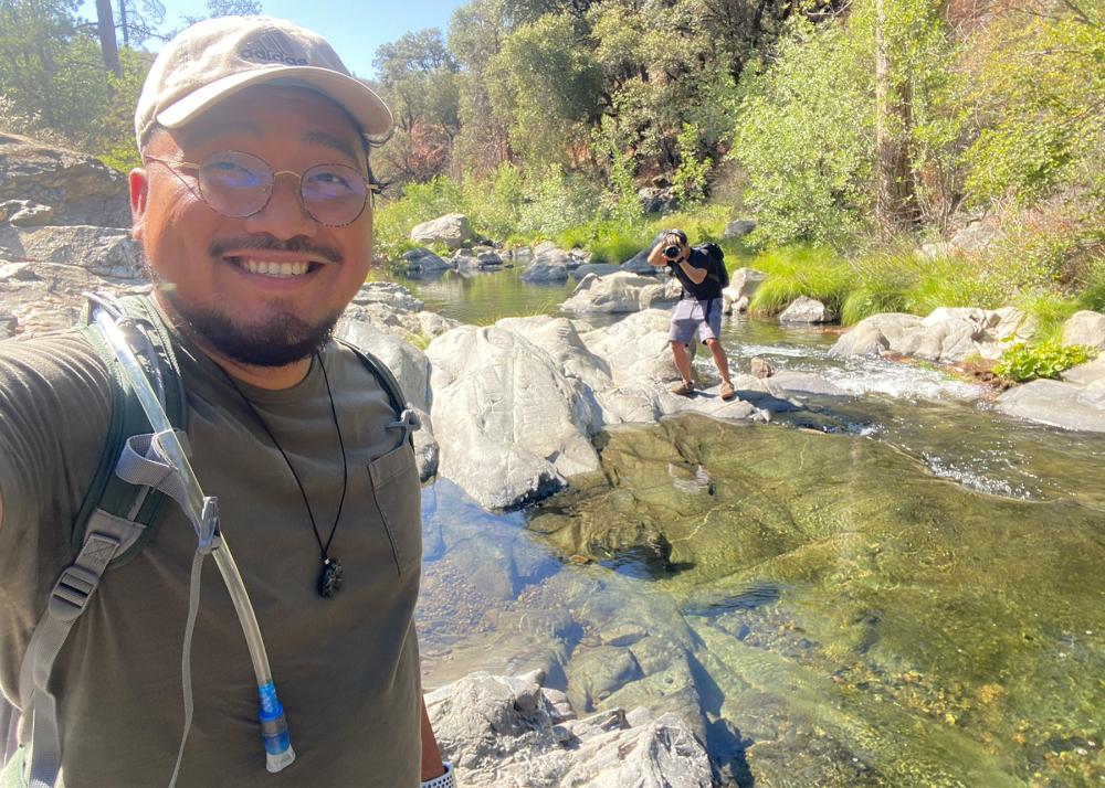 person taking a selfie, smiling, while another person is in the background crossing a river and posing for the photo