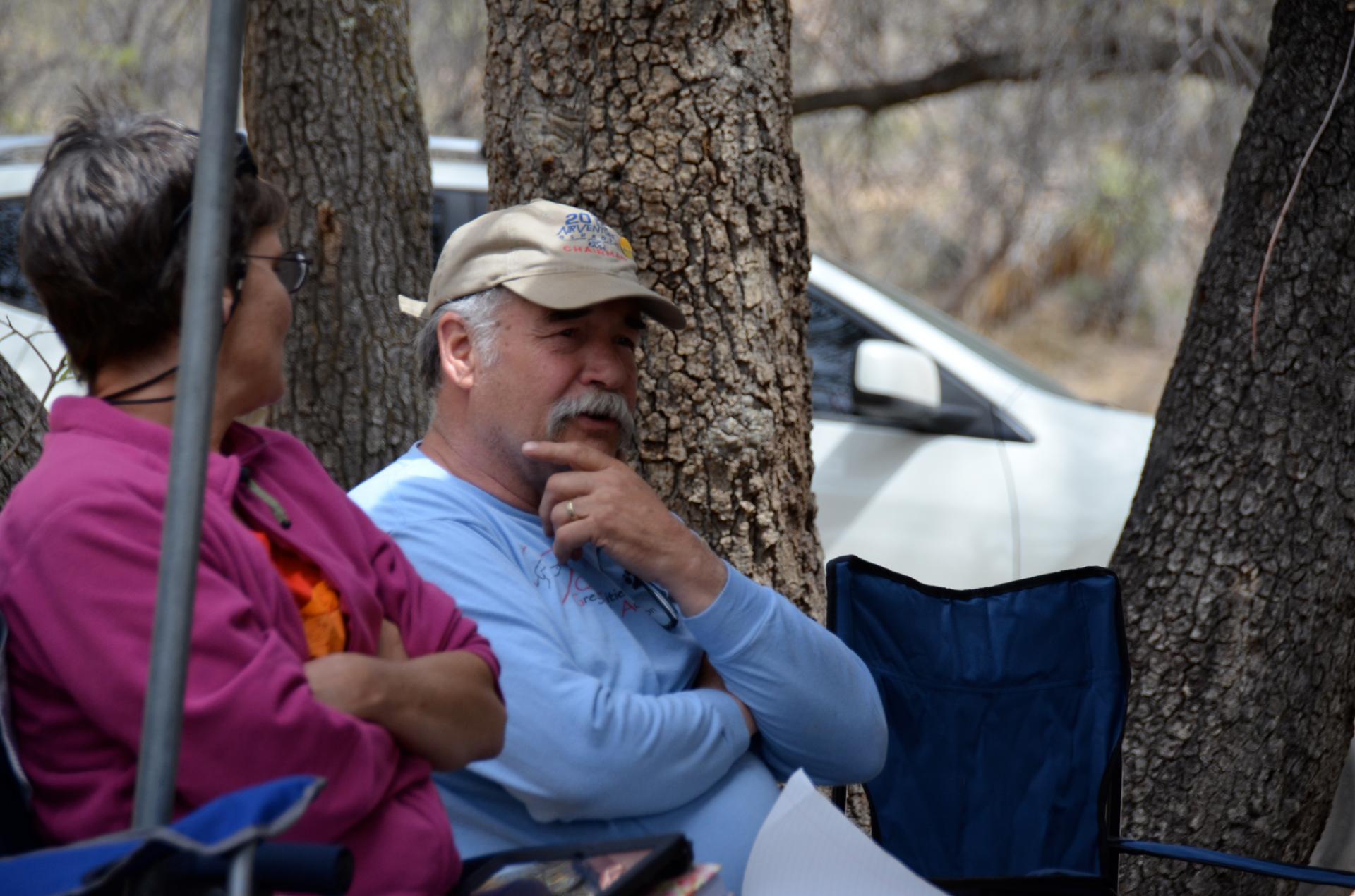 Man with gray mustache sits in front of trees and holds his hand to his chin