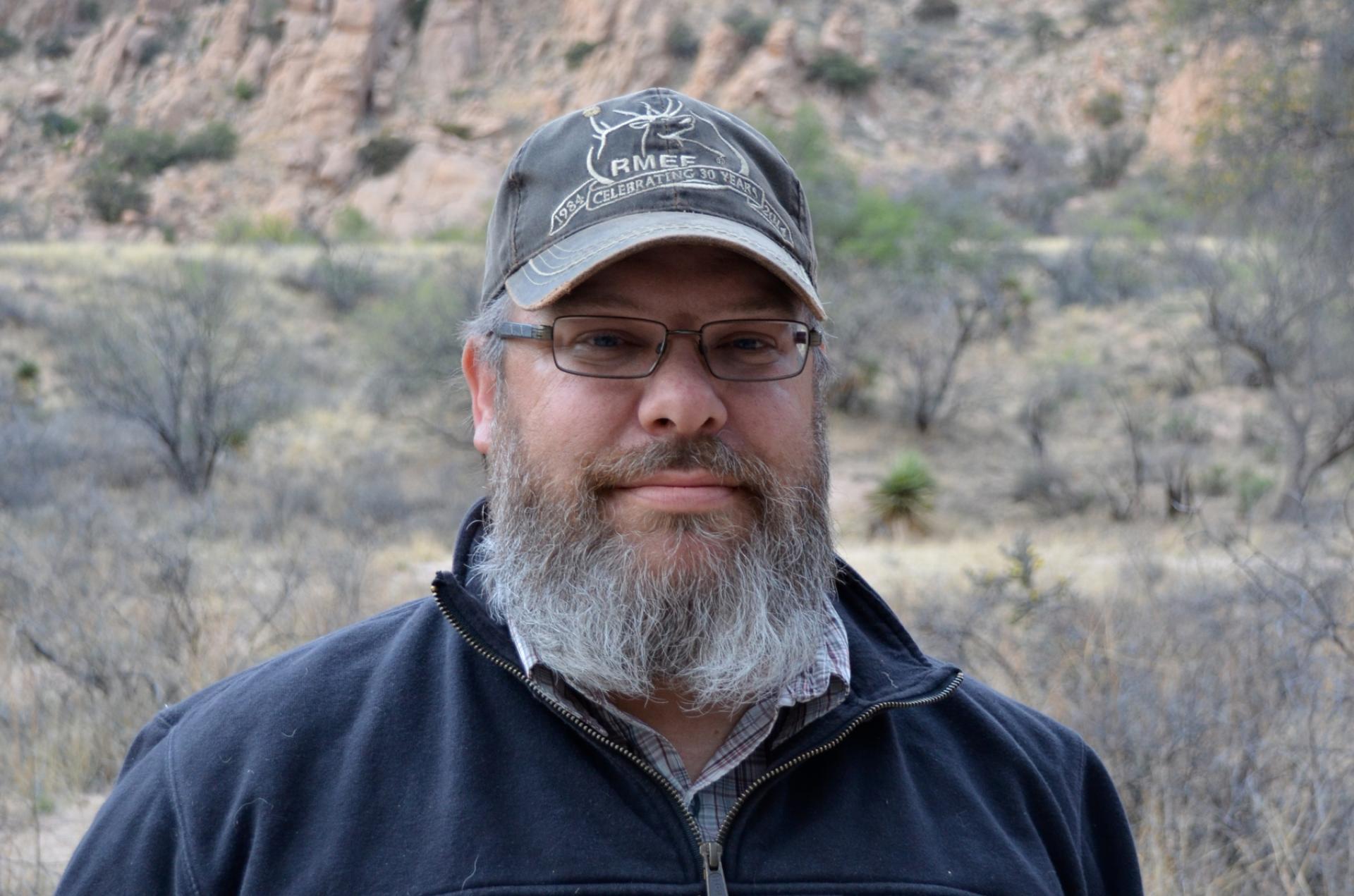 Man with gray beard, glasses and baseball cap standing in front of desert landscape