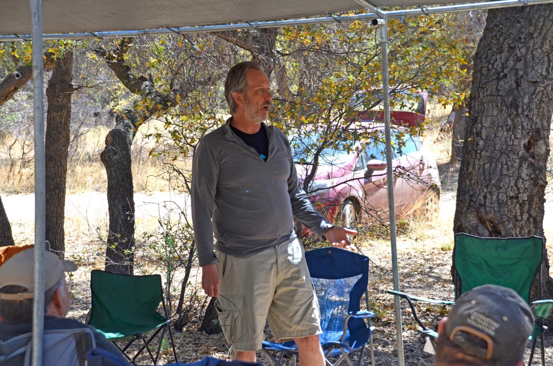 Man with gray beard standing and talking under canopy structure