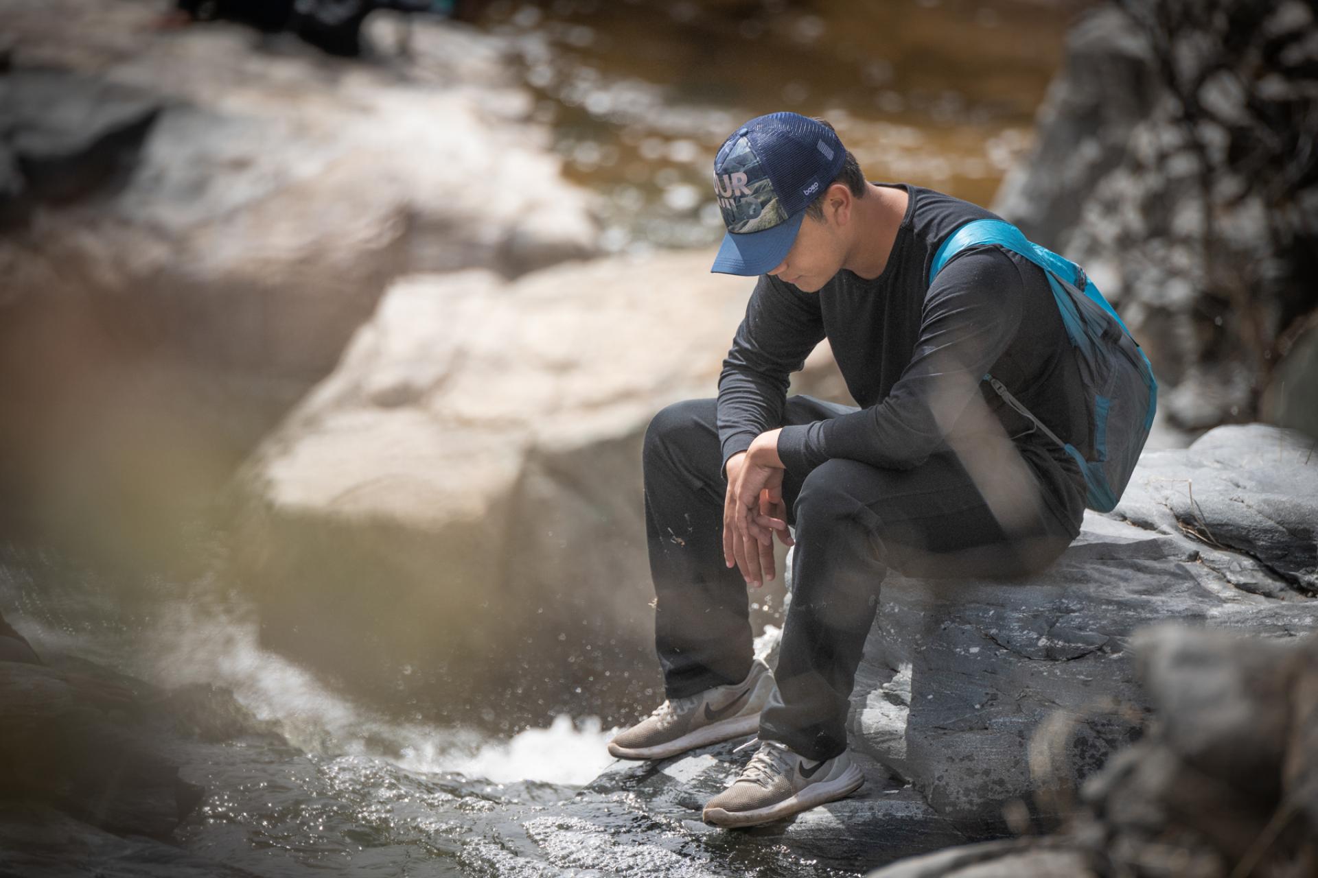 Person seated on rock next to creek, looking down thoughtfully with baseball cap on head and rocks in background
