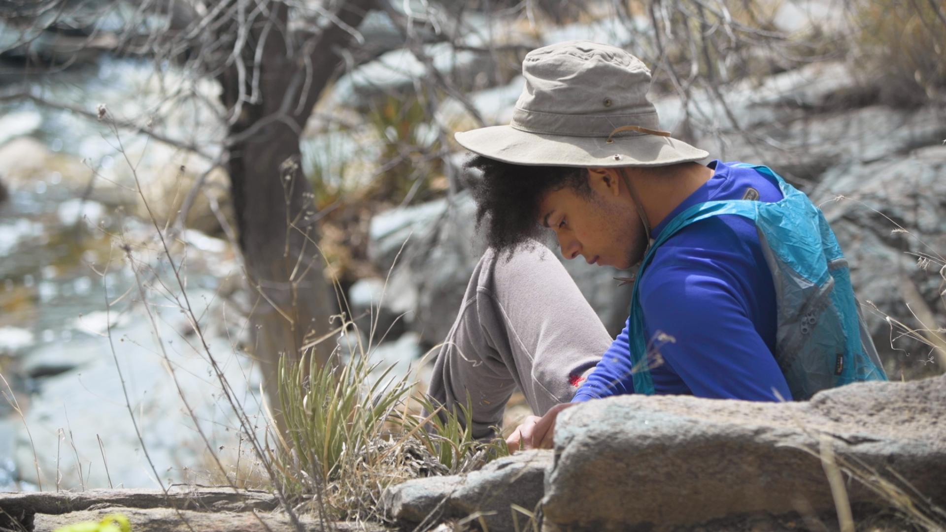 Person seated and wearing a brimmed hat, bent over and looking at something, with rocks and trees in the background