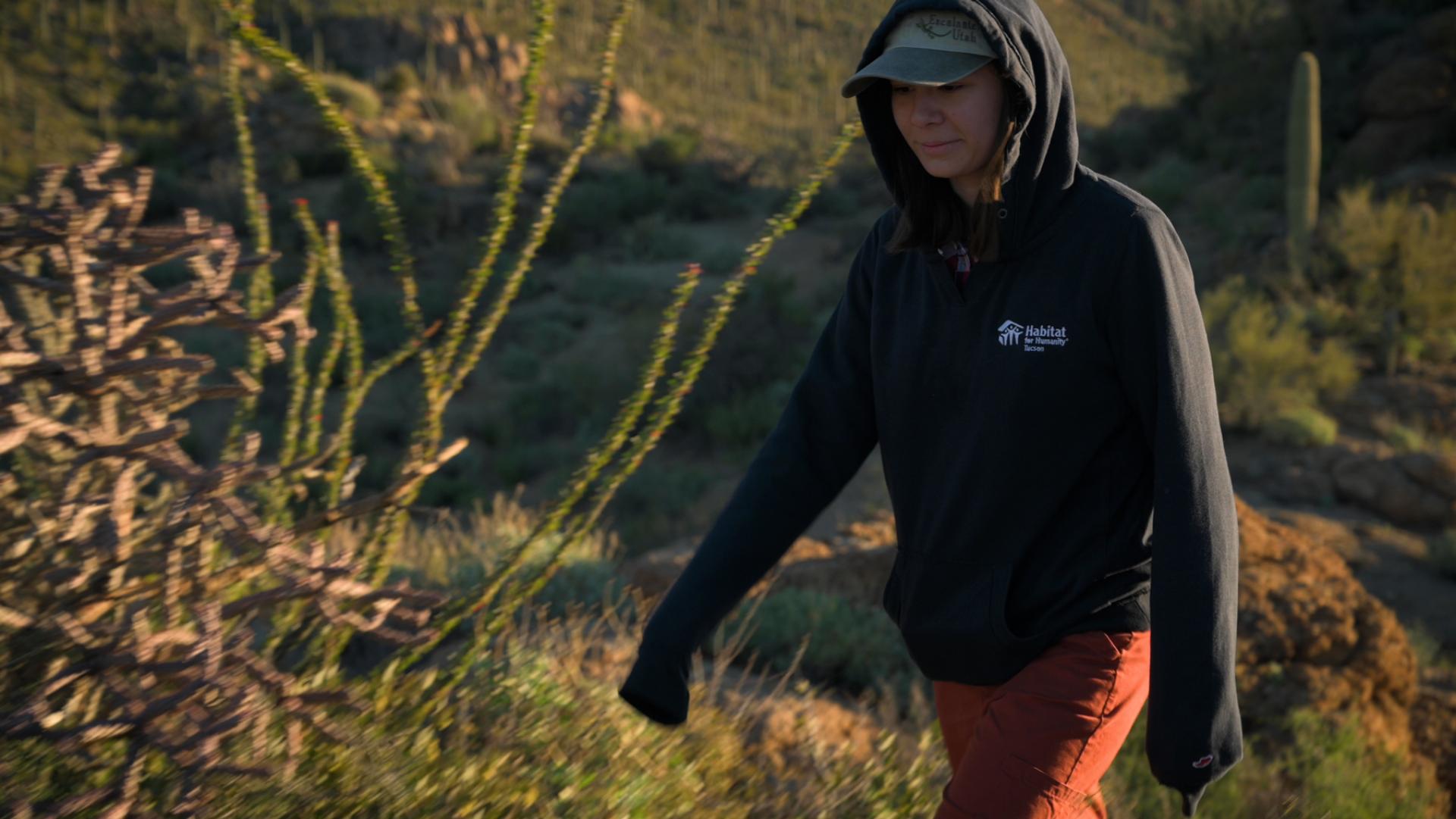 Person walking to the left wearing a dark hooded sweatshirt and baseball cap with desert plants in the background