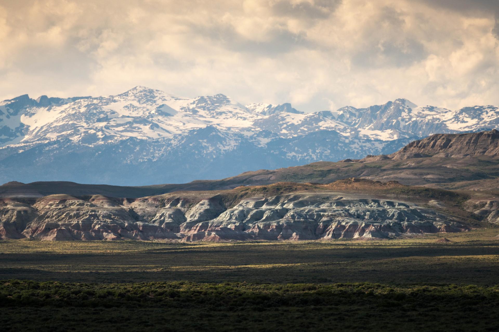 Snow capped mountains rising behind sandstone badlands and sagebrush under a cloudy sky in the Northern Red Desert, Wyoming