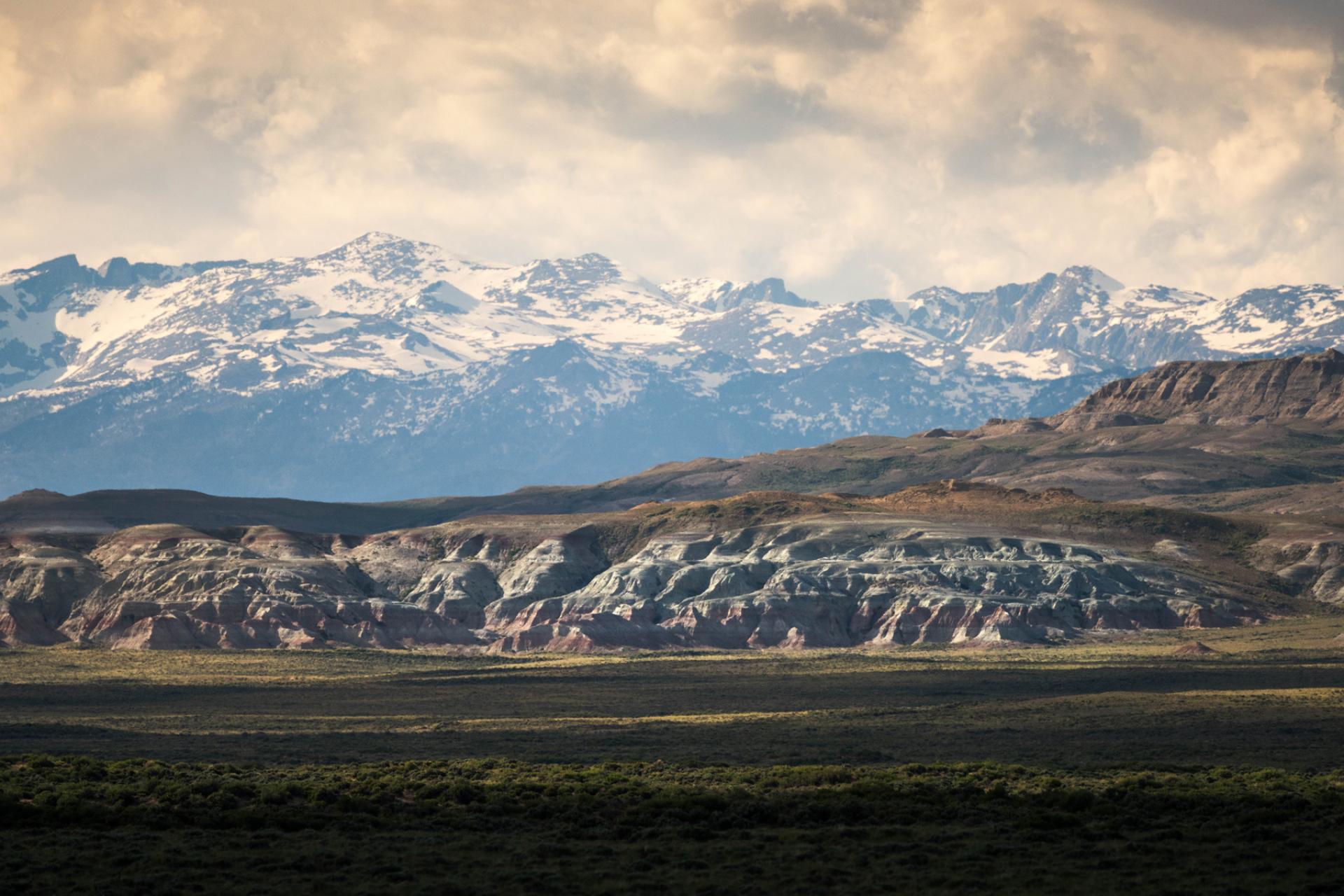 Snow capped mountains rising behind sandstone badlands and sagebrush under a cloudy sky in the Northern Red Desert, Wyoming
