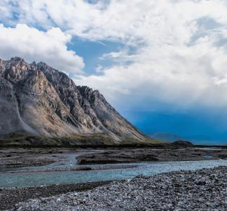 Cloudy skies above mountains and a river bordered by coarse rocky beaches