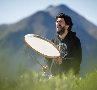 Bearded man using a stick to beat the face of a large, shallow drum with mountains in the background