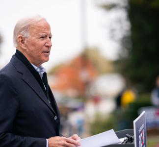 Joe Biden looking to the right and holding papers with trees and buildings in the background