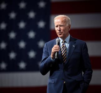 President Joe Biden holding a microphone and standing in front of a large American flag backdrop