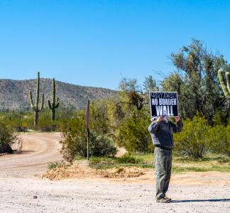 Person standing with sign that reads "No Border Wall" underneath blue sky with green cactus and other plants behind them