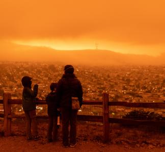Three people standing in front of a fence looking out over a cityscape with an all-encompassing hazy orange light overwhelming the scene
