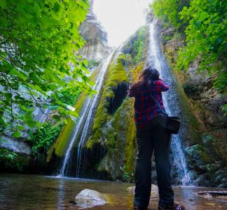 Person pictured from rear looking up and taking picture of waterfall surrounded by greenery