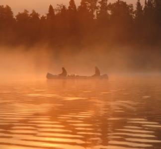 Two people in a canoe on a lake, partly obscured by haze in an orange partly sunlit scene
