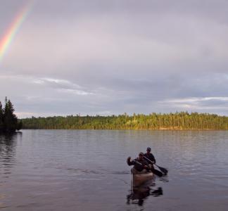 Two people in a canoe paddling toward foreground in a lake with a rainbow in the upper left corner, Boundary Waters Canoe Area Wilderness, Minnesota