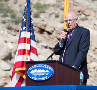 Man standing at lectern with microphone in his hand and an American flag in the background