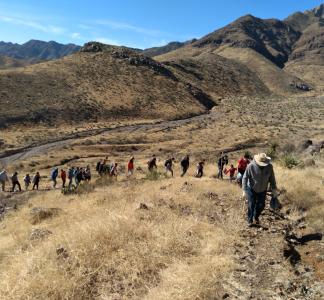 Procession of people hiking through mountainous desert landscape, with leader of the group near the foreground