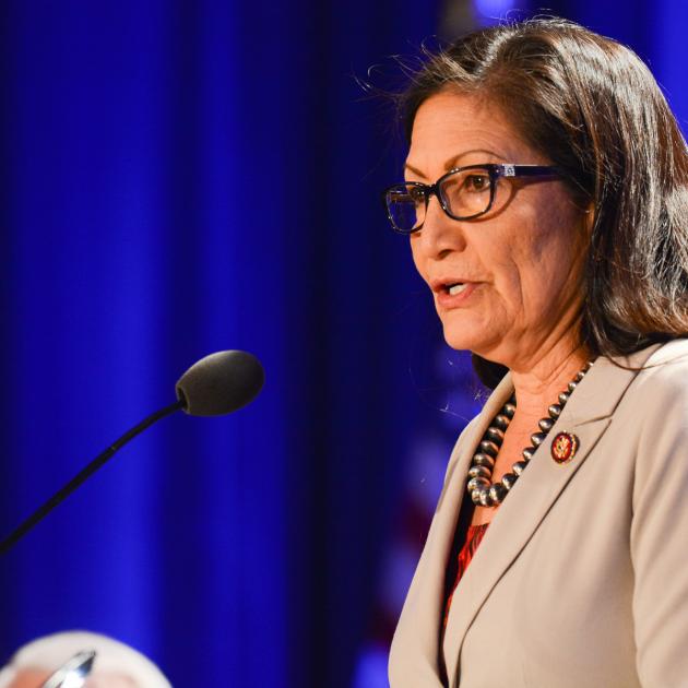 Woman with glasses speaking into microphone in front of blue curtain