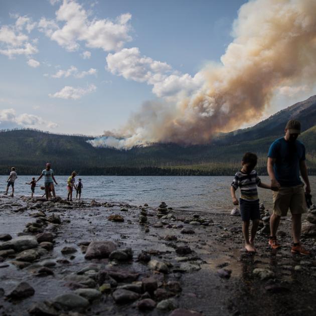 People walking along the edge of a lake in the foreground with forested landscape and a tall column of smoke visible in the distance