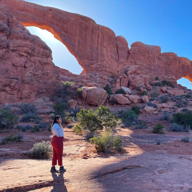 Woman standing on sandstone with trees in the back and a large stone wall with two openings in it against the blue sky