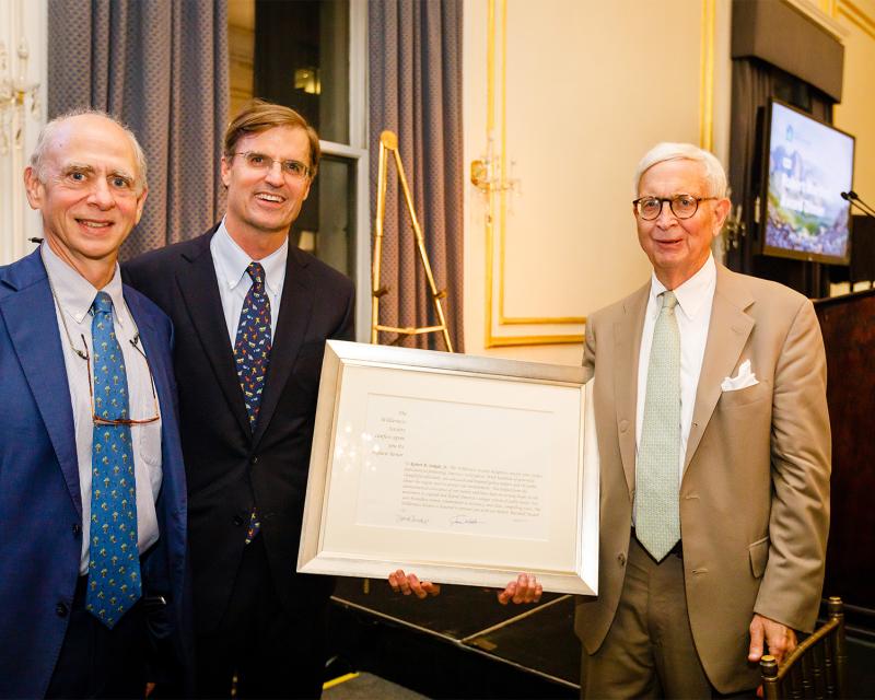 Three men in suits stand smiling with one of them holding a certificate 