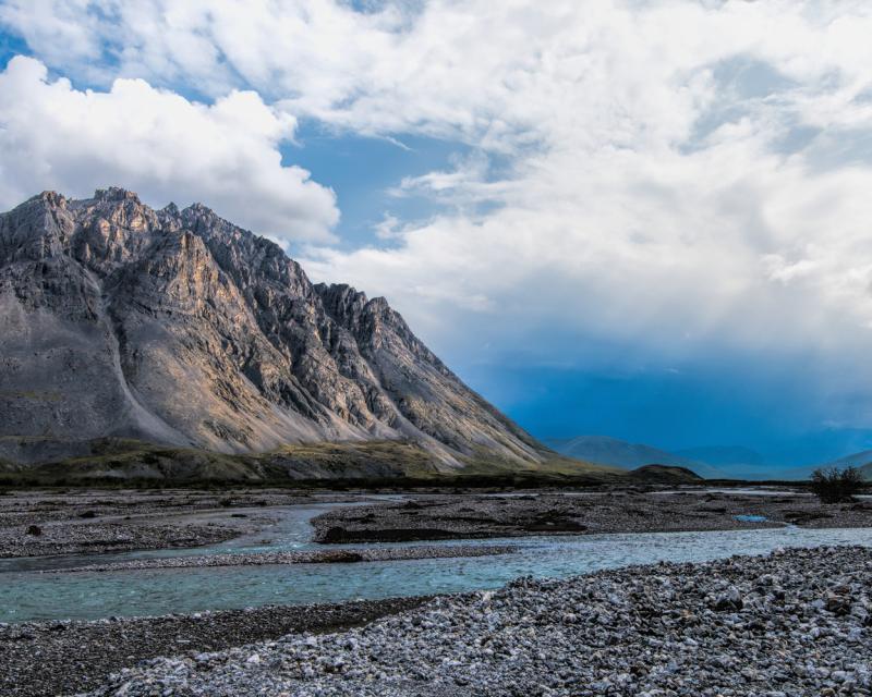 Cloudy skies above mountains and a river bordered by coarse rocky beaches