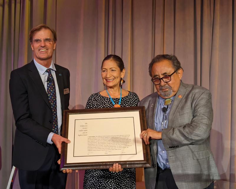 Rep. Raul Grijalva, Interior Secretary Deb Haaland and Wilderness Society President Jamie Williams standing next to each other in front of a curtain, holding a large framed document