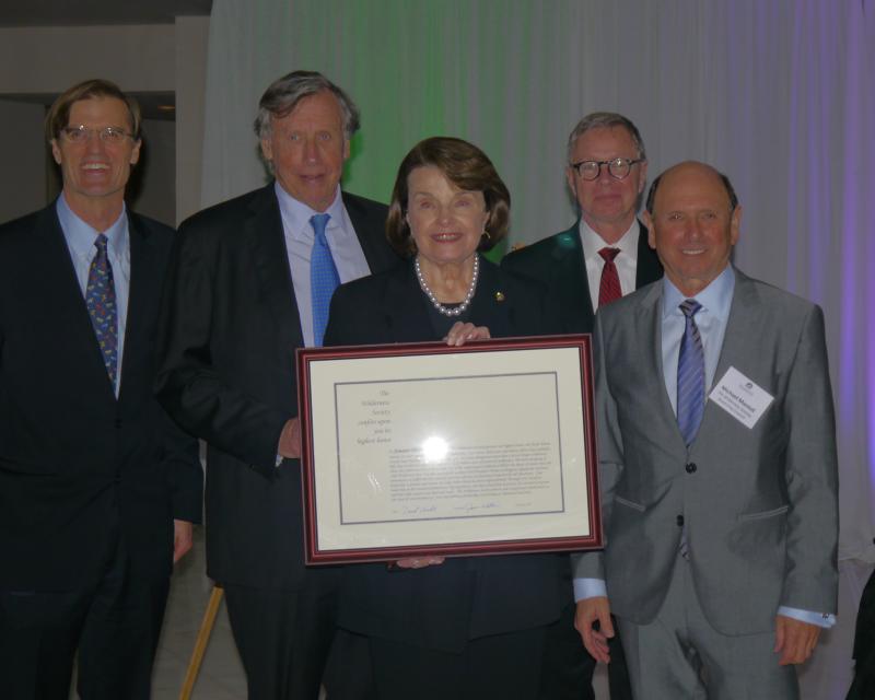 four men wearing suits surround a woman in the middle holding a plaque