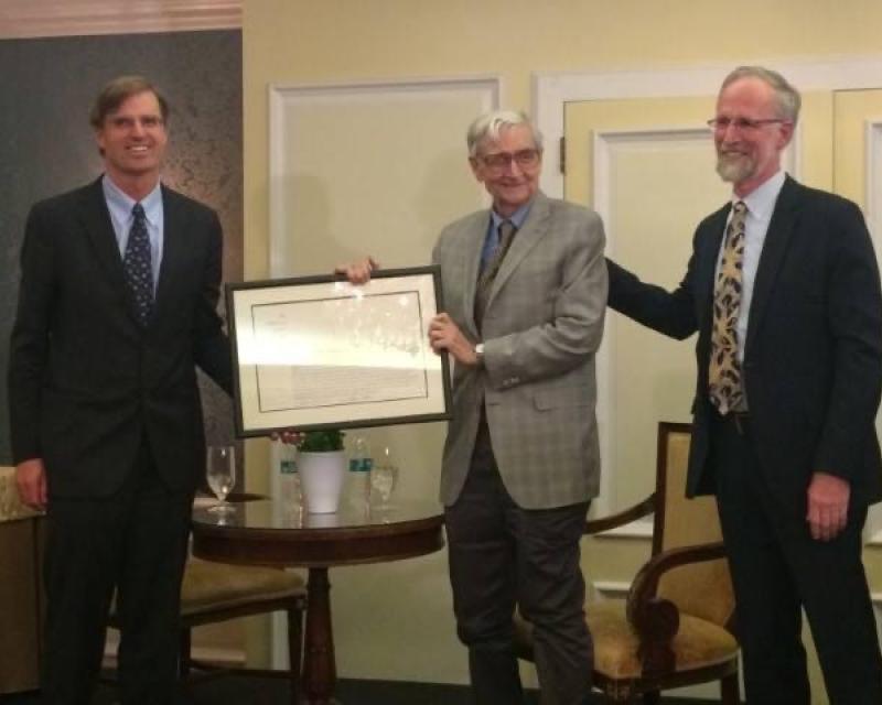 three men with suits stand next to each other with one of them holding a certificate